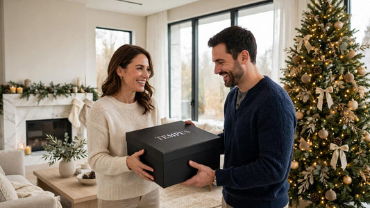 Wife handing her husband a black Tempus gift box beside the Christmas tree in a bright luxury living room